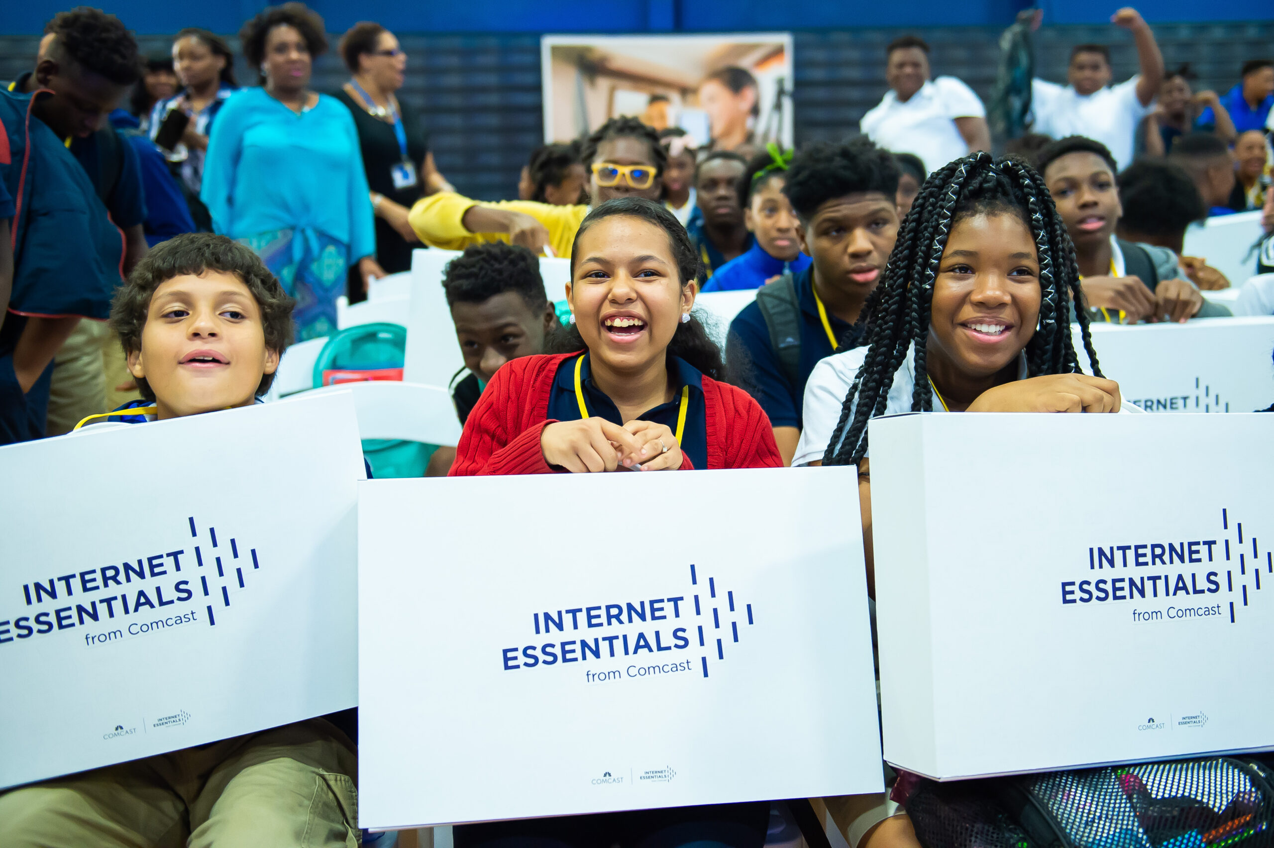 Students sit smiling holding boxes in their laps that say "Internet Essentials." Comcast Internet Essentials Program, an initiative to help bridge the digital divide among students.